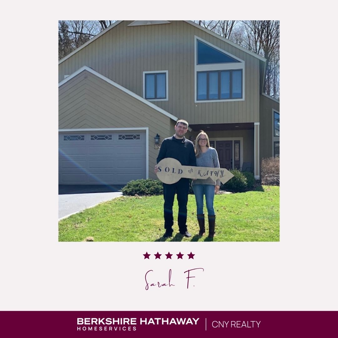 Two people in front of a light brown house holding a large key-shaped sign that says "sold by Kathy"