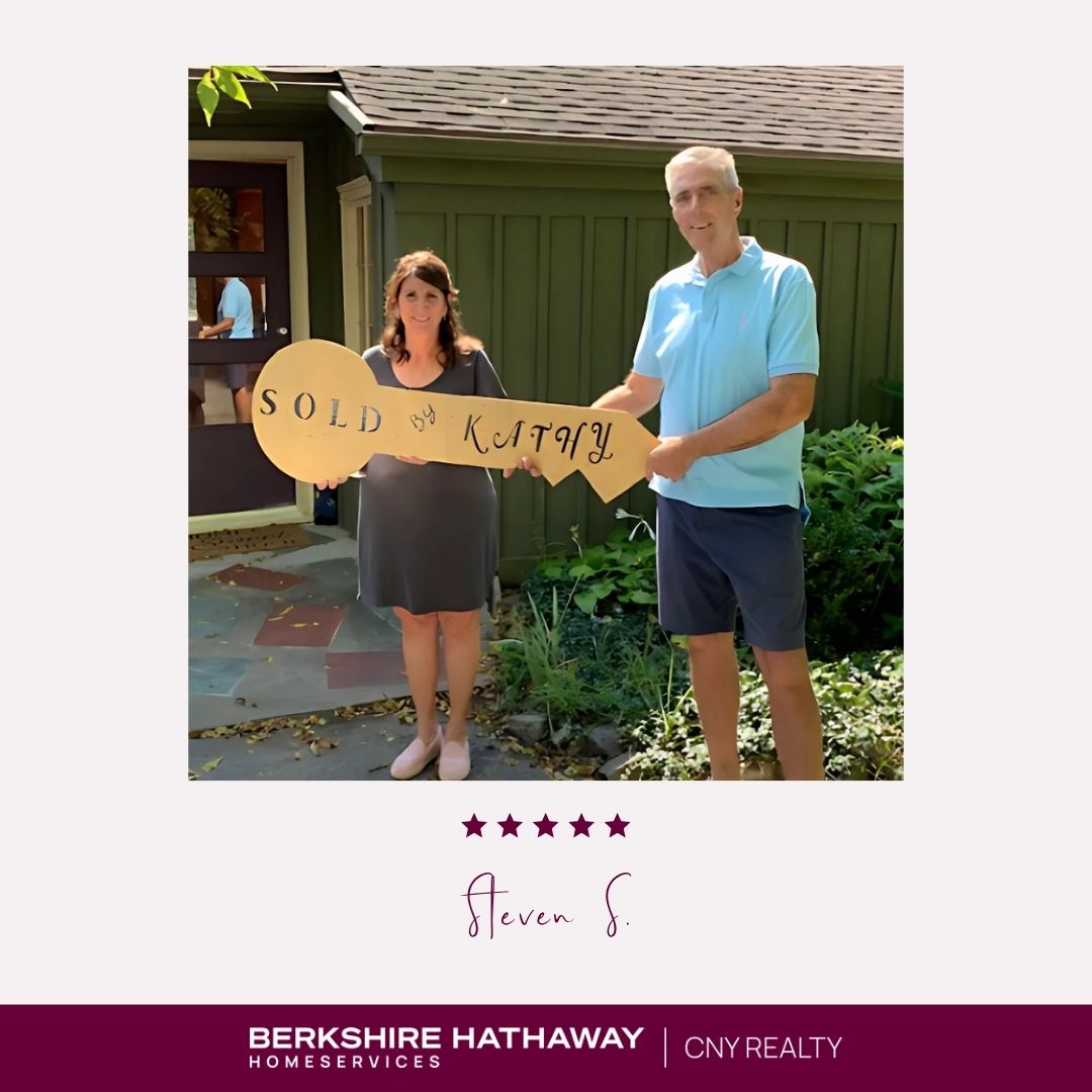 A man and a woman in front of a green house holding a large key-shaped sign that says "sold with Kathy."