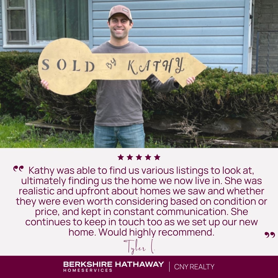 A person in front of a blue house holding a large key-shaped sign that says "Sold by Kathy"